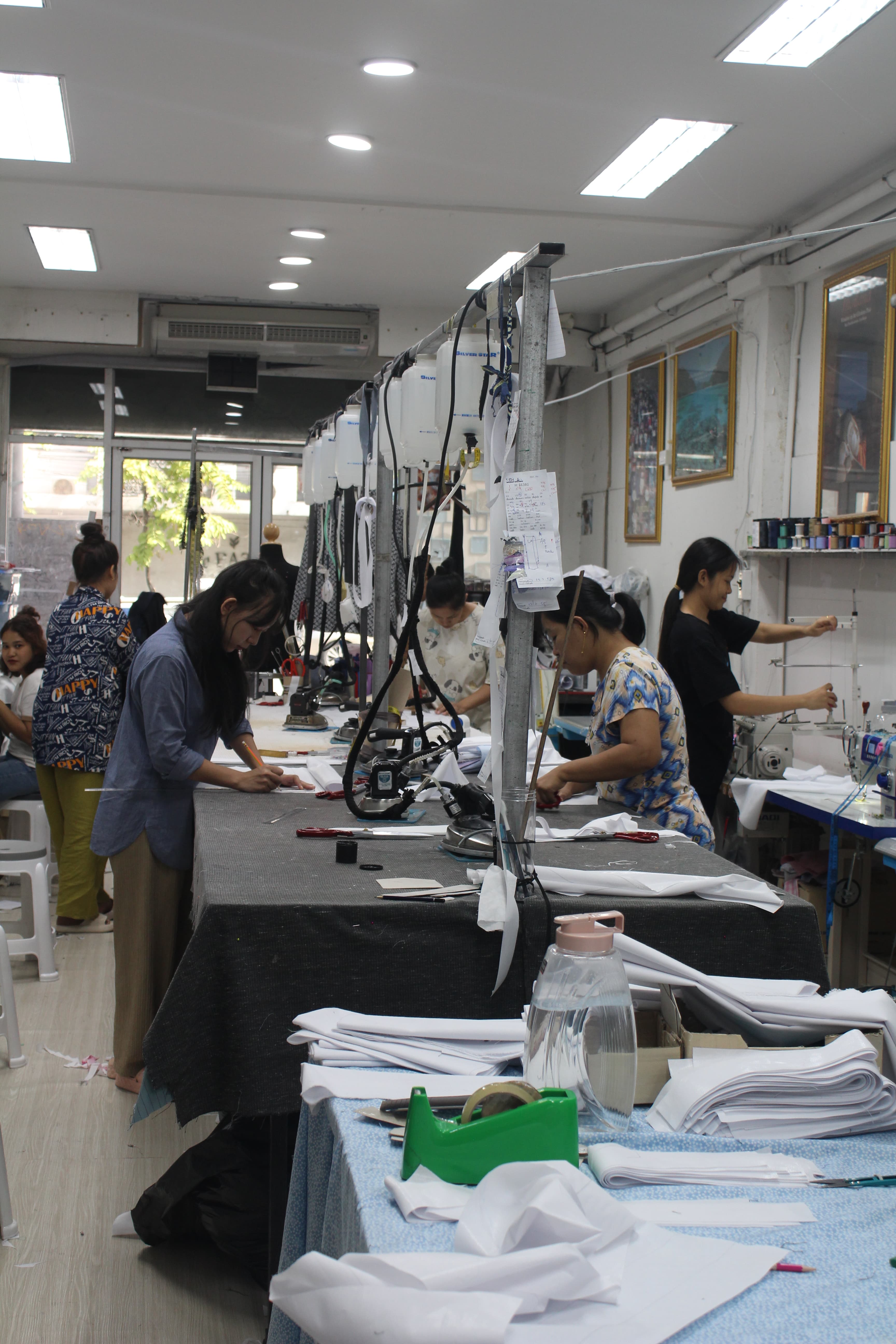 A bright workroom with sewing machines, paper patterns on the wall and a long table with a roll of grey cloth